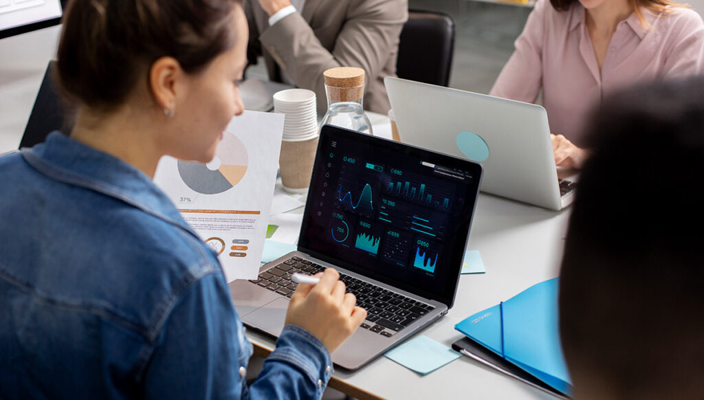 Affärsmöte där en kvinna analyserar finansiella data på en laptop med avancerade diagram, vilket visar hur AI projektarbete kan integreras i daglig verksamhet.