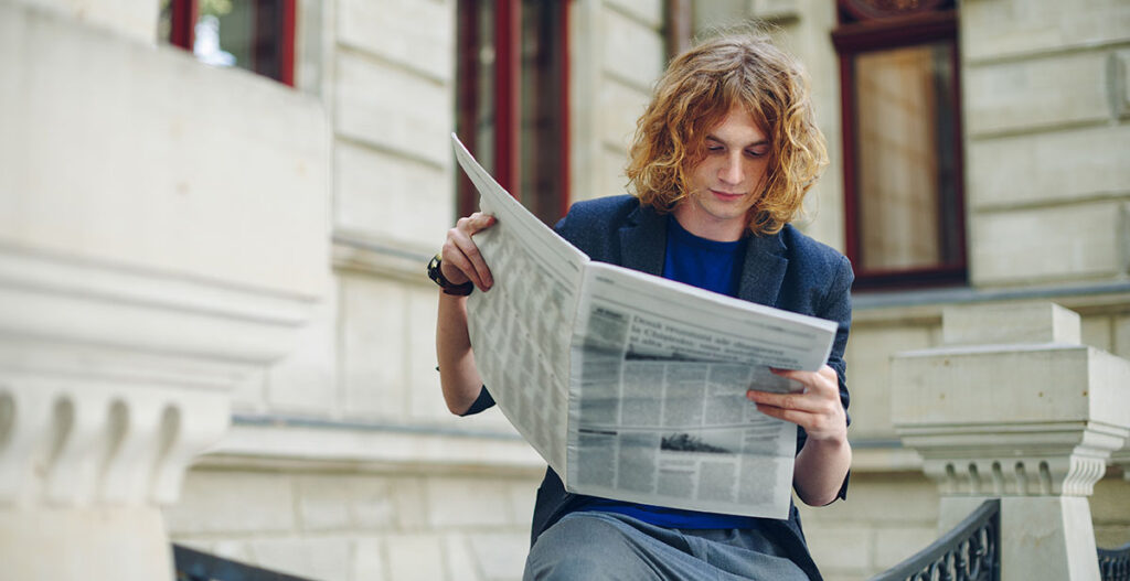 Ung akademiker läser tidning utanför en byggnad – illustrerar stigande arbetslöshet bland nyexaminerade amerikaner i en arbetsmarknad förändrad av AI